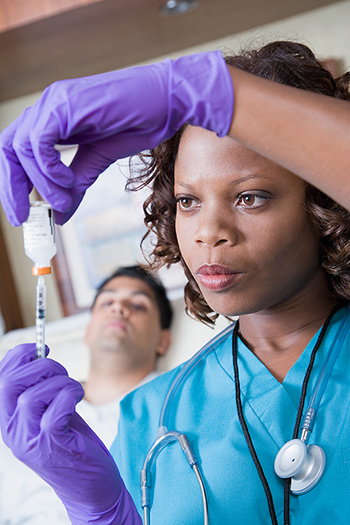 Nurse loading a needle with patient in hospital bed in background.