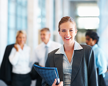 A professional woman smiles while holding a notebook with others in the background.